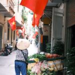 A street vendor pushes a cart filled with vegetables under Vietnamese flags in a narrow Hanoi alley.